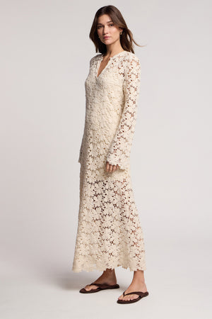 A woman models the JOVIE DRESS by Velvet by Graham & Spencer, a long sleeve lace maxi dress in cream crochet, styled with brown sandals against a plain light background.