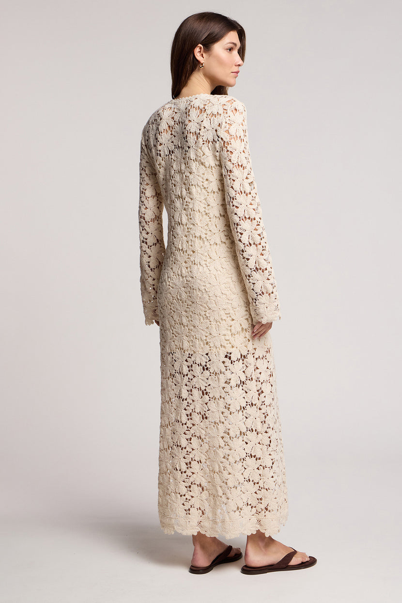 A woman stands in profile wearing the JOVIE DRESS by Velvet by Graham & Spencer, a cream long sleeve floral lace maxi with an ankle-length hem and flat brown sandals, set against a plain background.