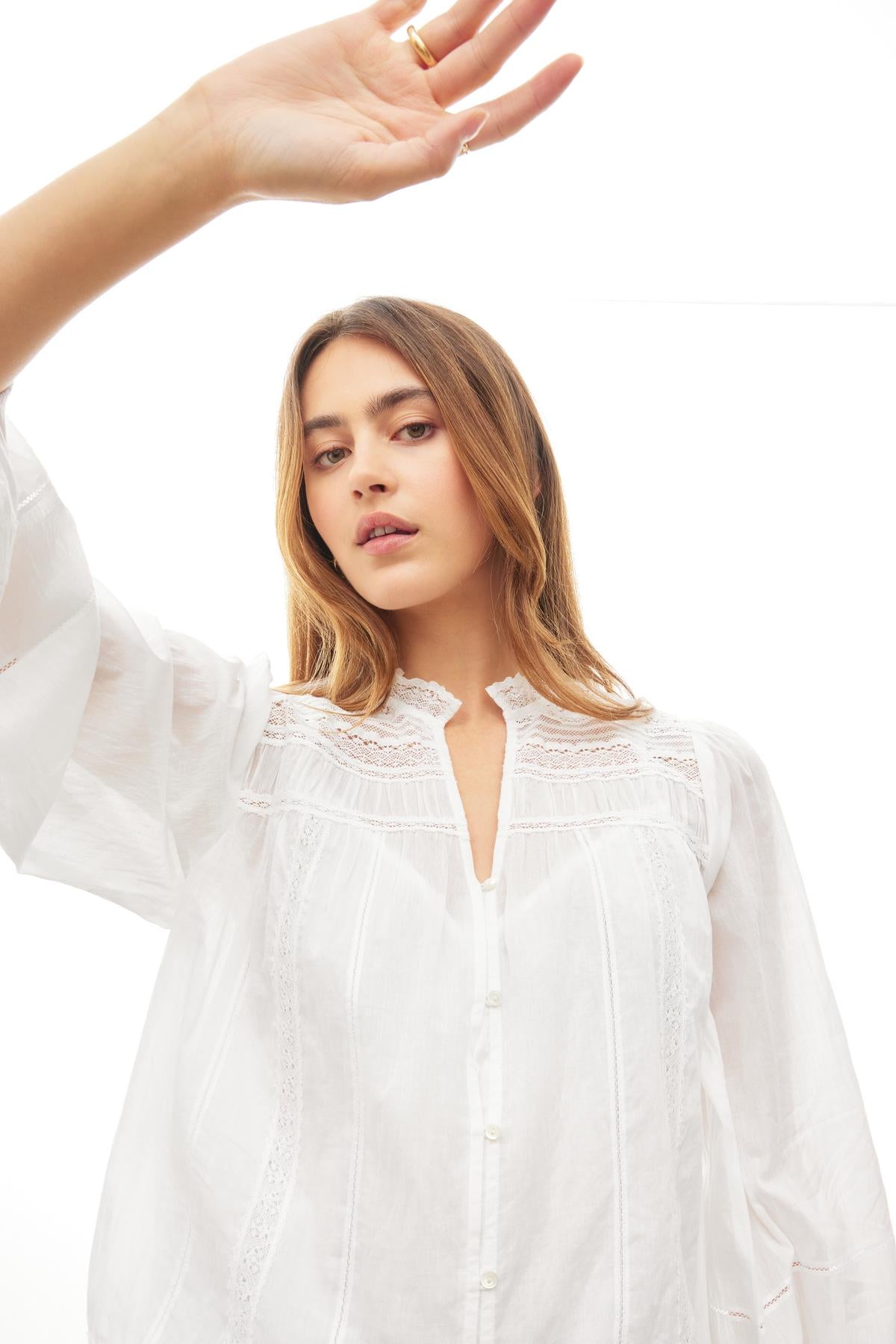   A person with long hair wears the Velvet by Graham & Spencer LIAM COTTON LACE BUTTON FRONT TOP with sheer sleeves and raises one hand, standing against a white background. 