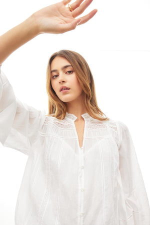 A person with long hair wears the Velvet by Graham & Spencer LIAM COTTON LACE BUTTON FRONT TOP with sheer sleeves and raises one hand, standing against a white background.