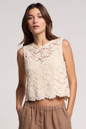 A woman wears the SIMONA TOP by Velvet by Graham & Spencer—a floral lace crop top with a scallop hem—paired with light brown pants, standing against a neutral background.