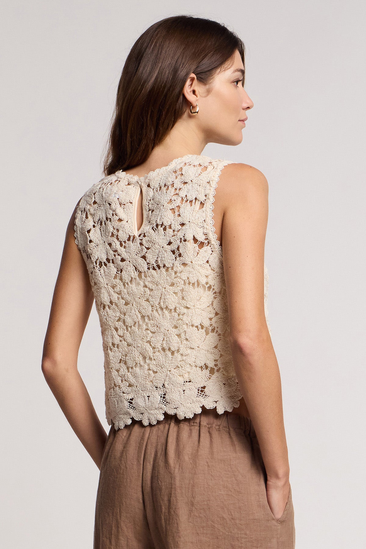 A person stands sideways in a cream floral lace crop top with scallop hem—SIMONA TOP by Velvet by Graham & Spencer—and light brown pants, against a plain light background.