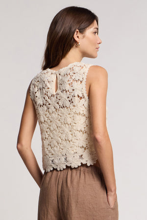 A person stands sideways in a cream floral lace crop top with scallop hem—SIMONA TOP by Velvet by Graham & Spencer—and light brown pants, against a plain light background.