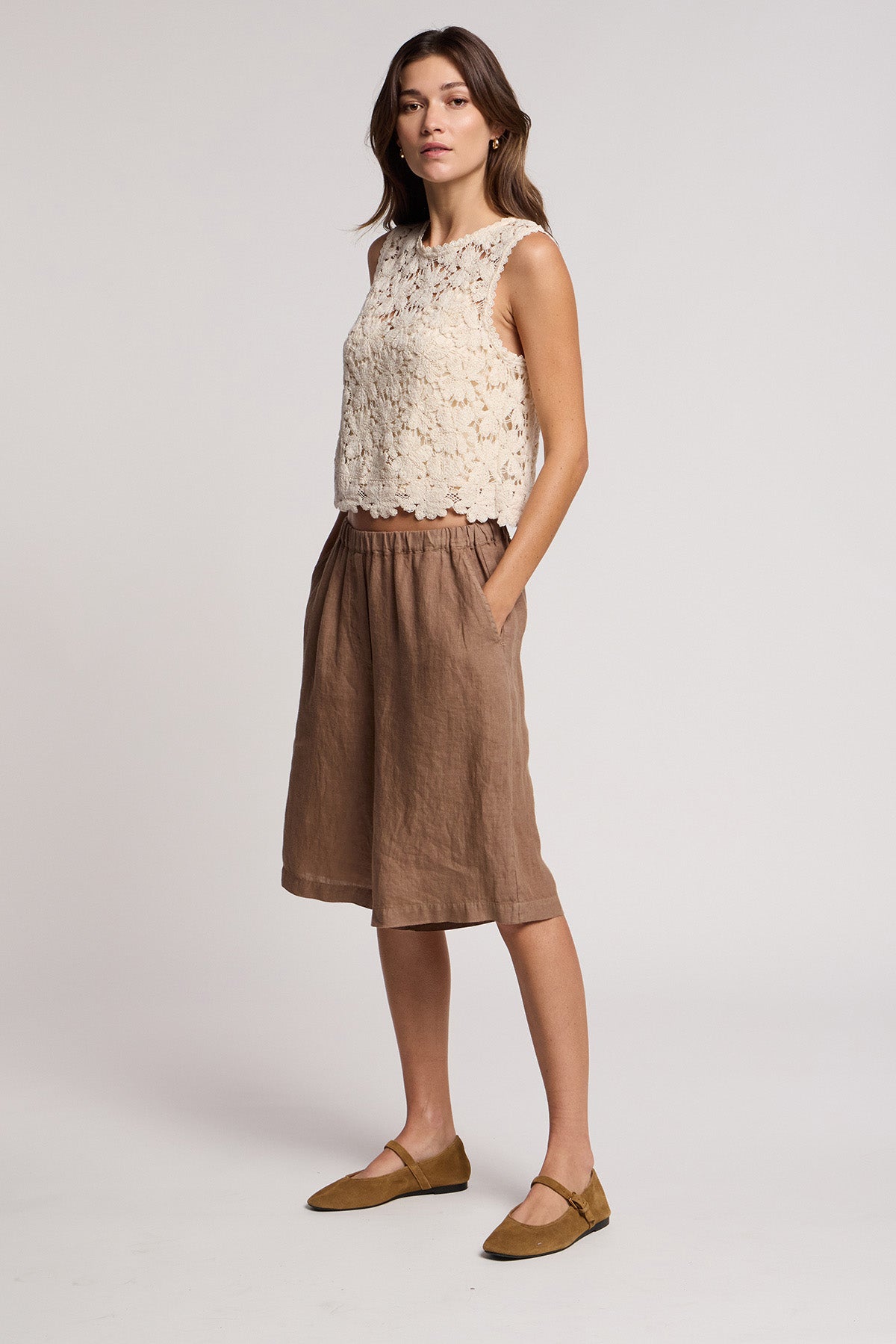 A woman wears the SIMONA TOP by Velvet by Graham & Spencer—a sleeveless cream floral lace crop top—paired with brown knee-length shorts and brown flats, standing against a plain background with her hands in her pockets.-41372217835713