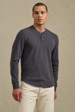 A man stands facing forward, wearing the ARNOLD HENLEY by Velvet by Graham & Spencer in dark gray and off-white pants against a plain beige background.