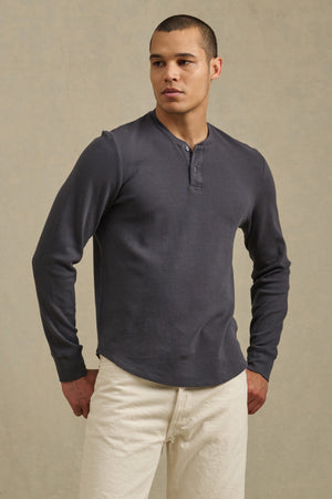 A person wearing the ARNOLD HENLEY by Velvet by Graham & Spencer in dark gray with light-colored pants stands against a neutral background.