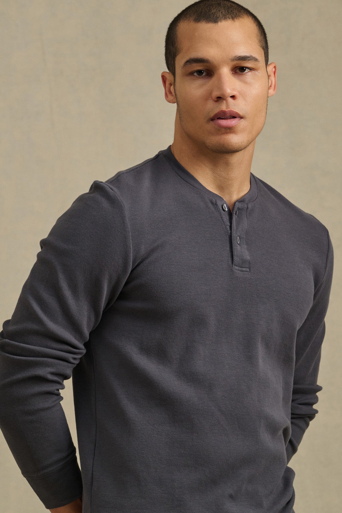 Someone wearing the ARNOLD HENLEY by Velvet by Graham & Spencer, a long-sleeve dark gray shirt, stands facing the camera with a neutral expression against a plain beige background.