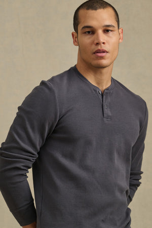 Someone wearing the ARNOLD HENLEY by Velvet by Graham & Spencer, a long-sleeve dark gray shirt, stands facing the camera with a neutral expression against a plain beige background.