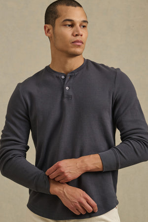 A man stands against a beige background with his arms crossed, wearing the Velvet by Graham & Spencer ARNOLD HENLEY, a long-sleeve dark gray shirt with a two-button placket, and looks to the side.