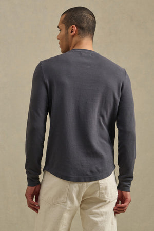 A person faces away, wearing the ARNOLD HENLEY long-sleeve dark gray shirt by Velvet by Graham & Spencer and light-colored pants against a plain, neutral background.