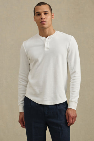 A man stands facing forward in the ARNOLD HENLEY by Velvet by Graham & Spencer, a white long-sleeve shirt, paired with dark pants against a plain beige background.