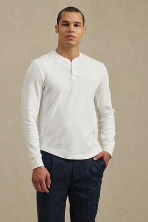 A man stands against a plain backdrop, wearing the ARNOLD HENLEY by Velvet by Graham & Spencer—a white long-sleeve henley shirt—paired with dark pants, one hand in his pocket.