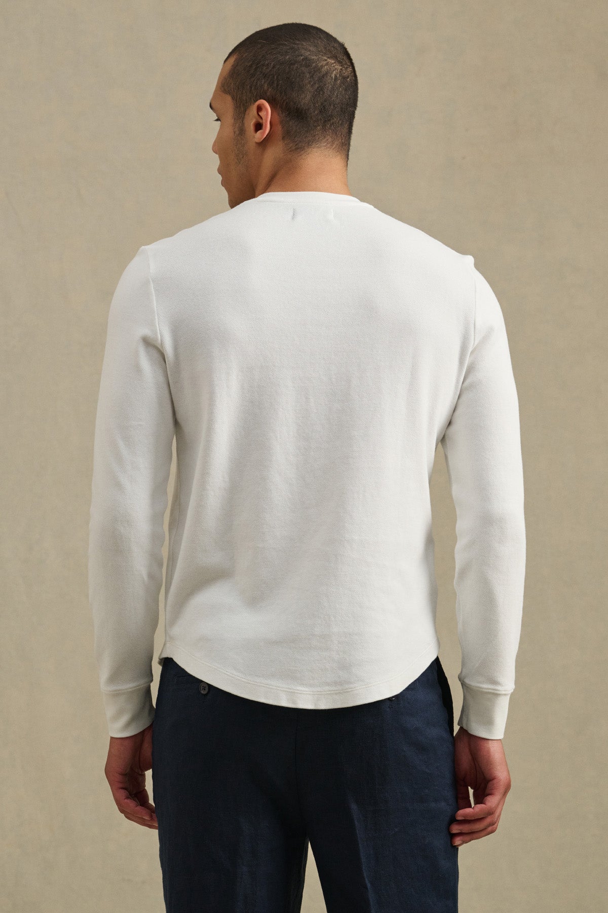 A man faces away from the camera, wearing the ARNOLD HENLEY by Velvet by Graham & Spencer—a white long-sleeve shirt paired with dark pants—set against a neutral background.-41374344773825