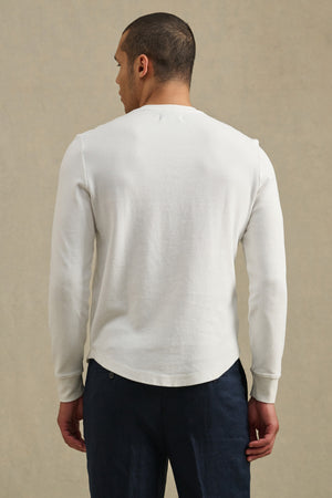 A man faces away from the camera, wearing the ARNOLD HENLEY by Velvet by Graham & Spencer—a white long-sleeve shirt paired with dark pants—set against a neutral background.