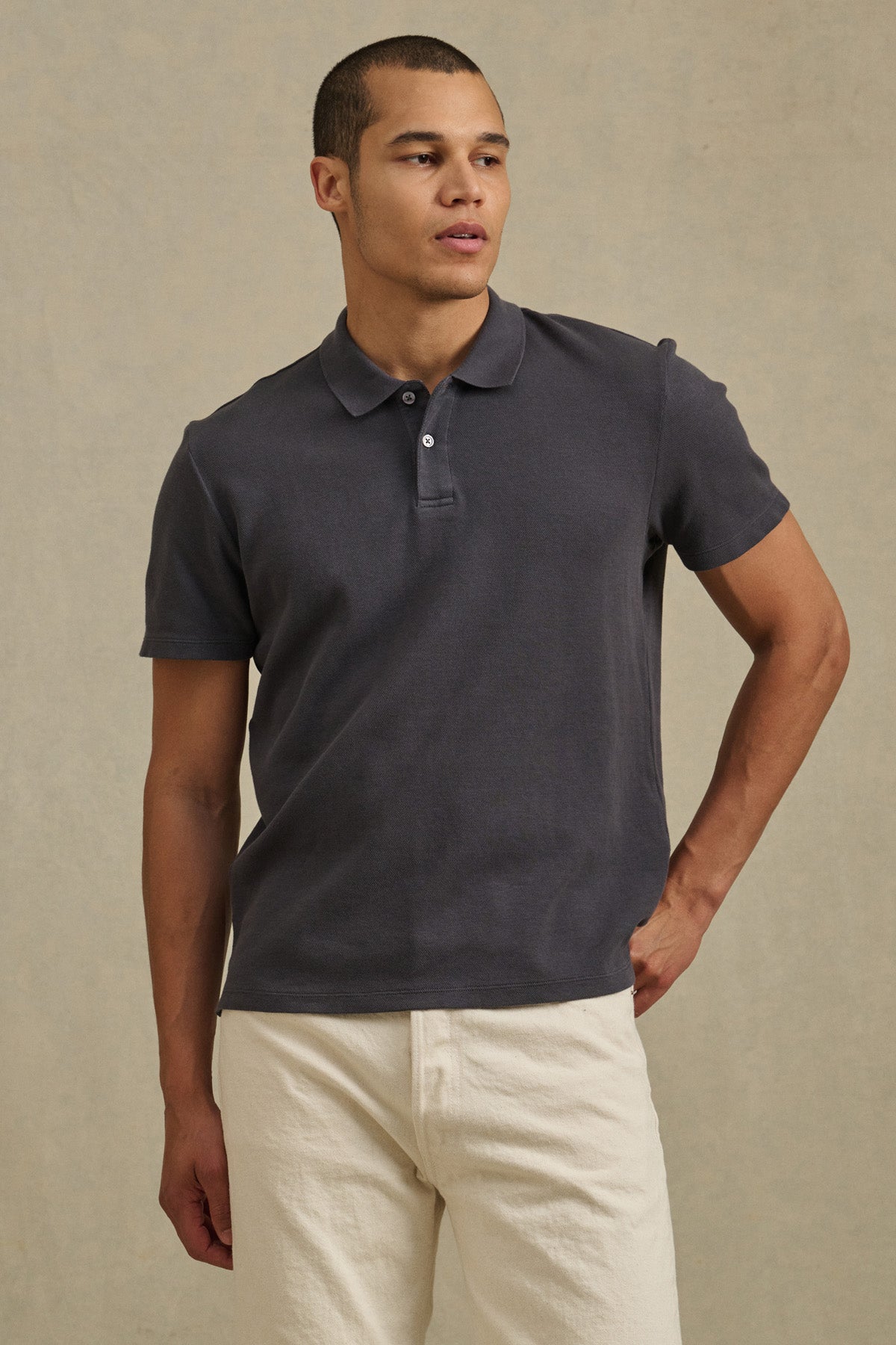 A man poses against a plain background wearing the dark gray JOHNNY POLO from Velvet by Graham & Spencer and light-colored pants, with one hand on his hip.