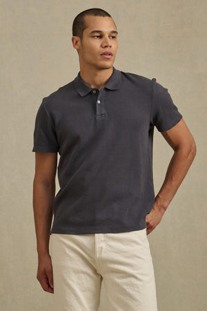 A man poses against a plain background wearing the dark gray JOHNNY POLO from Velvet by Graham & Spencer and light-colored pants, with one hand on his hip.