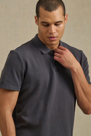 A man wearing the JOHNNY POLO by Velvet by Graham & Spencer in dark grey stands against a neutral background, looking down and touching his collar.