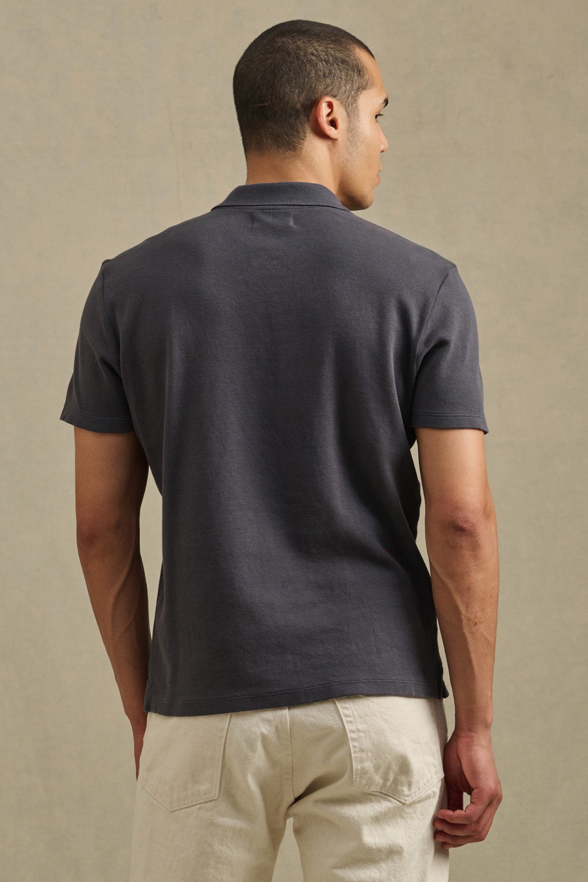 A man faces away, wearing the JOHNNY POLO short-sleeve dark gray shirt by Velvet by Graham & Spencer and off-white pants, set against a neutral background.