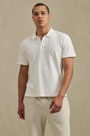 A man stands against a neutral background wearing the JOHNNY POLO by Velvet by Graham & Spencer, a white short-sleeve cotton mesh polo shirt, paired with light-colored pants.
