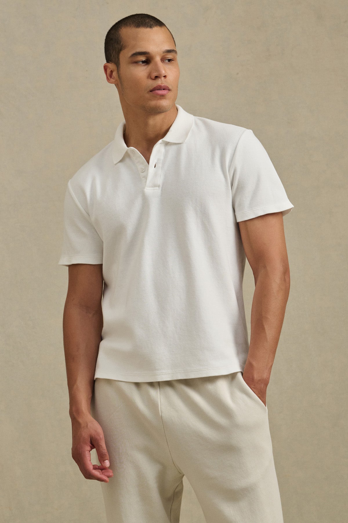 A man stands against a neutral background wearing the JOHNNY POLO by Velvet by Graham & Spencer and off-white pants, looking slightly to the side with one hand in his pocket.