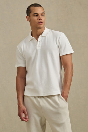 A man stands against a neutral background wearing the JOHNNY POLO by Velvet by Graham & Spencer and off-white pants, looking slightly to the side with one hand in his pocket.