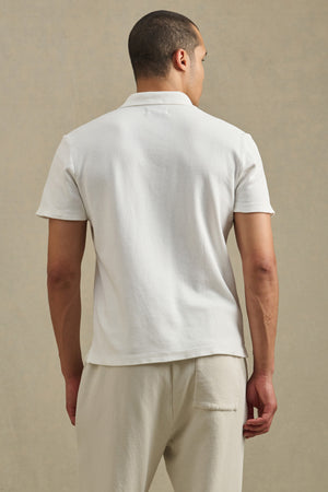 A man faces away from the camera in the Velvet by Graham & Spencer JOHNNY POLO, a breathable cotton mesh shirt, paired with light pants against a neutral background.