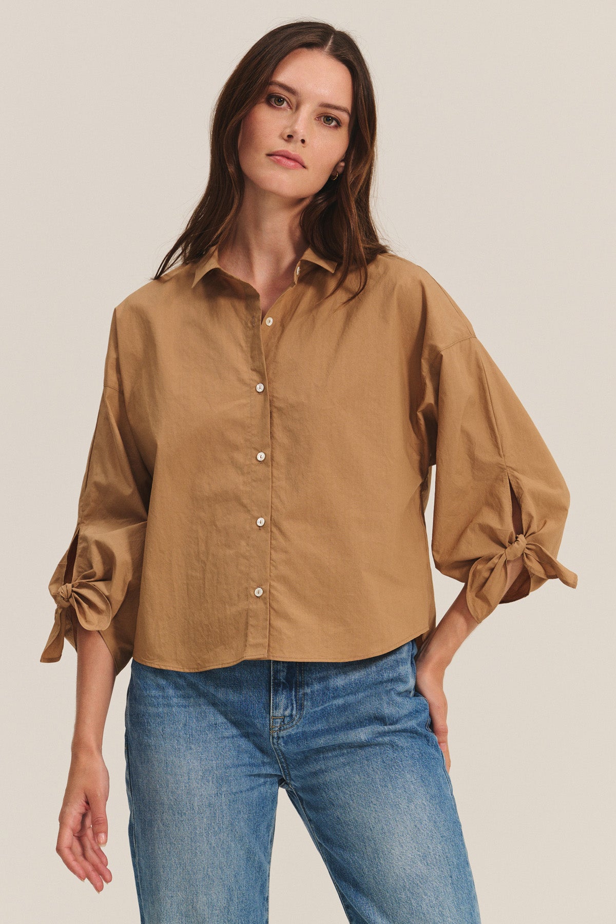A woman wears the ARLETTE SHIRT by Velvet by Graham & Spencer—a loose tan cotton poplin top with a classic collar and bow sleeve details—paired with blue jeans against a plain background.