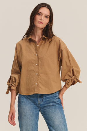 A woman wears the ARLETTE SHIRT by Velvet by Graham & Spencer—a loose tan cotton poplin top with a classic collar and bow sleeve details—paired with blue jeans against a plain background.