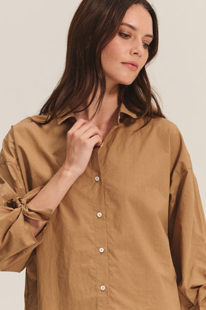 A woman in the ARLETTE SHIRT by Velvet by Graham & Spencer, a loose tan cotton poplin top with a classic collar and rolled-up sleeves, gazes downward with a neutral expression.