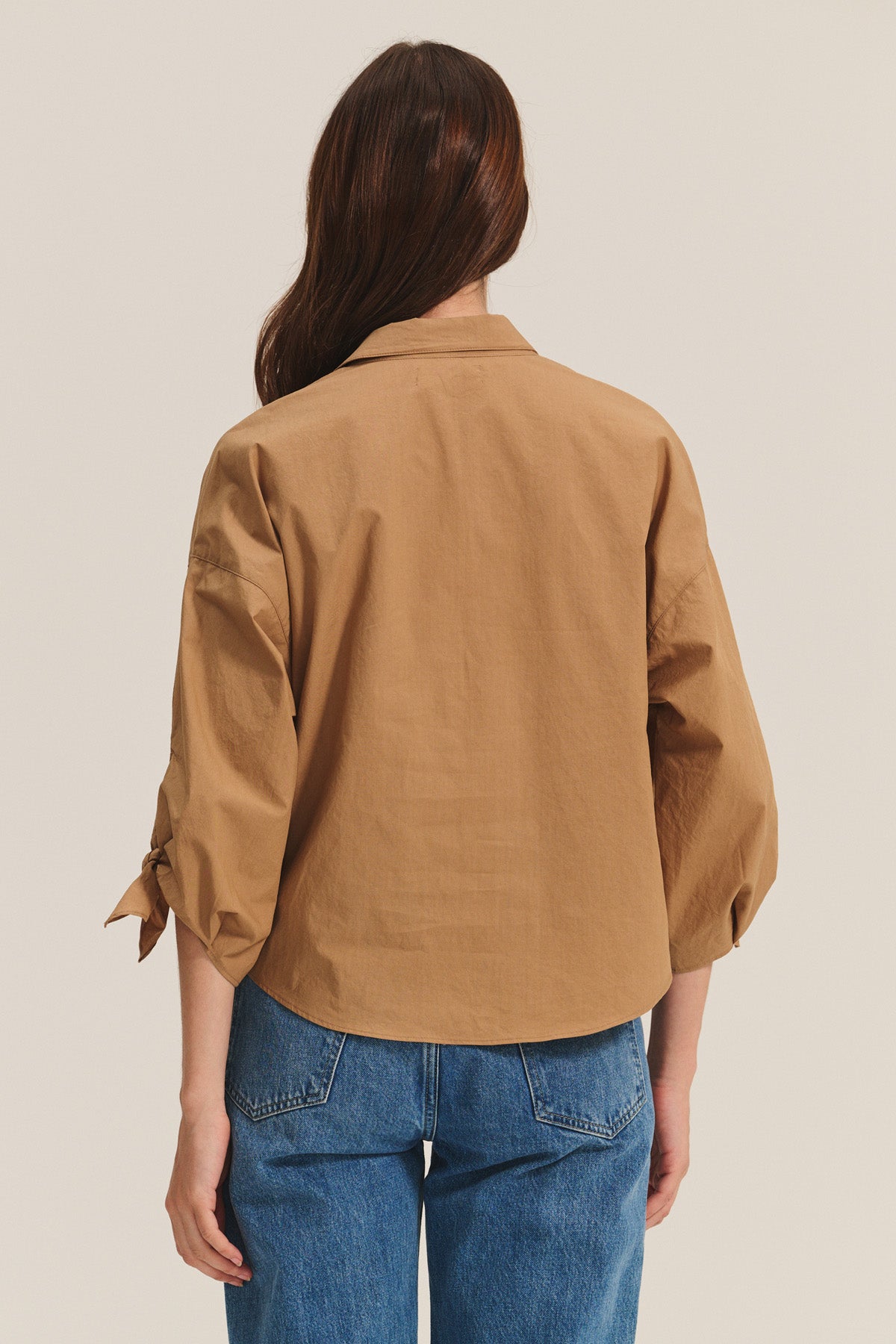 A person with long brown hair is seen from the back wearing the ARLETTE SHIRT by Velvet by Graham & Spencer—a tan cotton poplin top—paired with blue jeans, set against a plain light background.