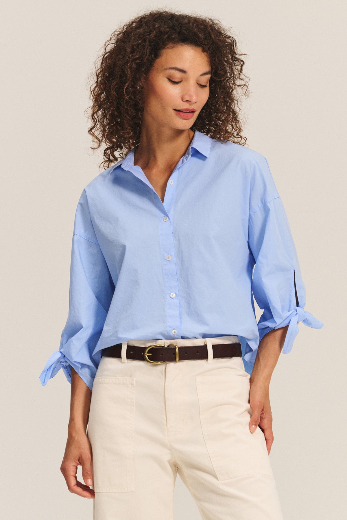 A woman wears the ARLETTE SHIRT by Velvet by Graham & Spencer—a light blue cotton poplin top with a classic collar and tied sleeves—paired with off-white pants, standing with her hand in her pocket against a plain background.-40333182337217