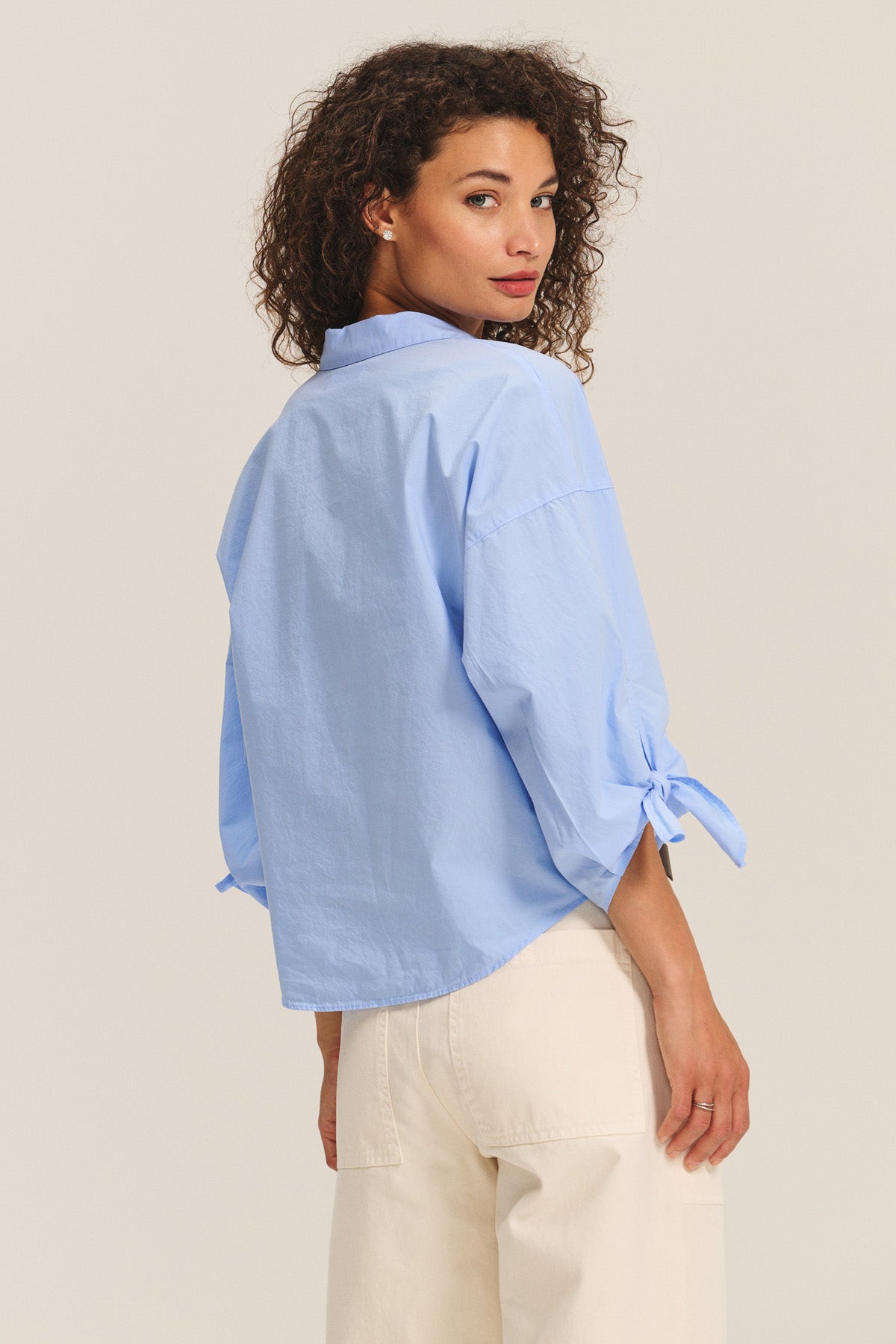 A woman with curly hair wears the ARLETTE SHIRT by Velvet by Graham & Spencer—a light blue cotton poplin top with a classic collar and tied cuffs—paired with off-white pants, standing and looking over her shoulder.-40333182304449