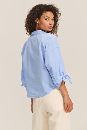 A woman with curly hair wears the ARLETTE SHIRT by Velvet by Graham & Spencer—a light blue cotton poplin top with a classic collar and tied cuffs—paired with off-white pants, standing and looking over her shoulder.