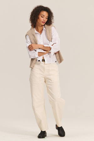 A woman stands with arms crossed, layering the Velvet by Graham & Spencer MAISIE SWEATER over a white button-up shirt, paired with cream pants and black shoes against a plain background.