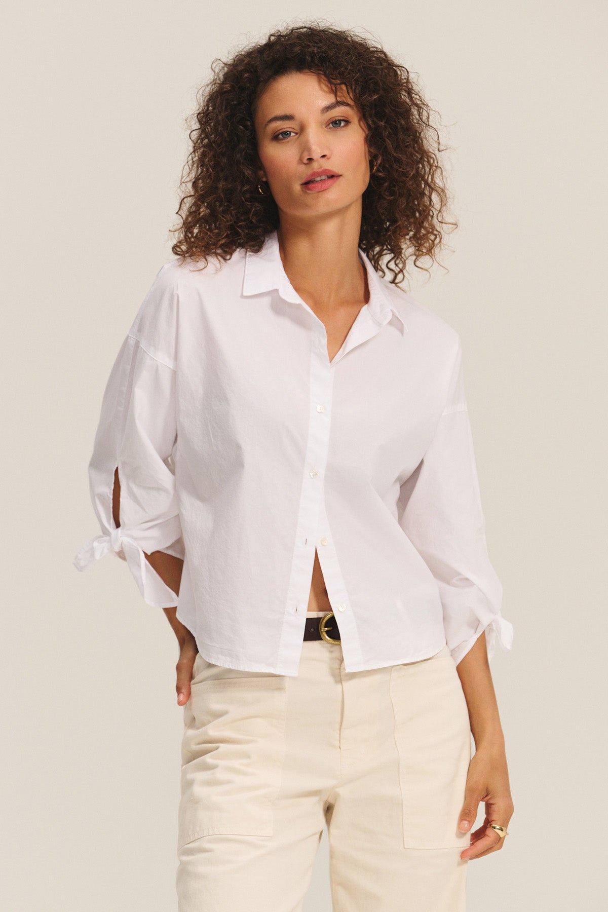 A woman wears the ARLETTE SHIRT by Velvet by Graham & Spencer—a cotton poplin top with a classic collar and decorative sleeve ties—paired with beige pants, standing against a plain light background.