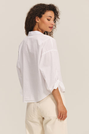 A person is shown from the back in a neutral setting, wearing the ARLETTE SHIRT by Velvet by Graham & Spencer—a loose white cotton poplin top with a classic collar and rolled-up sleeves—paired with off-white pants.