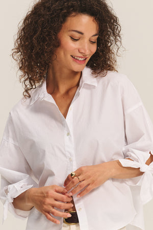 A woman with curly hair smiles wearing the ARLETTE SHIRT by Velvet by Graham & Spencer, a cotton poplin top with a classic collar and tied sleeves, paired with several rings on her fingers.