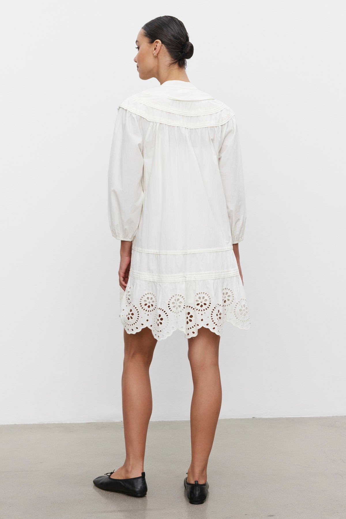   A person stands with their back facing the camera, wearing the BARB DRESS by Velvet by Graham & Spencer - a white, long-sleeve cotton dress featuring a lace hem, paired with black flat shoes. 