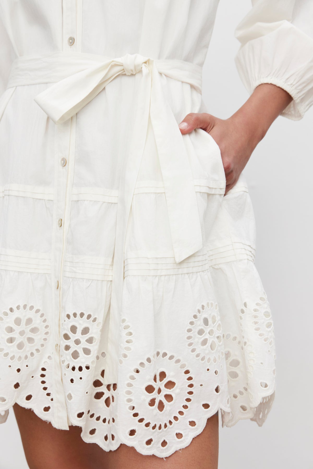   Close-up of a person wearing the BARB DRESS by Velvet by Graham & Spencer, featuring a white cotton fabric with broderie anglaise detailing and a waist tie. The customizable fit is accentuated by the pleated skirt, and the person has one hand tucked into a pocket. 