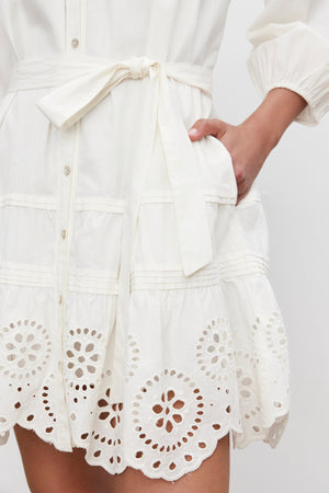 Close-up of a person wearing the BARB DRESS by Velvet by Graham & Spencer, featuring a white cotton fabric with broderie anglaise detailing and a waist tie. The customizable fit is accentuated by the pleated skirt, and the person has one hand tucked into a pocket.