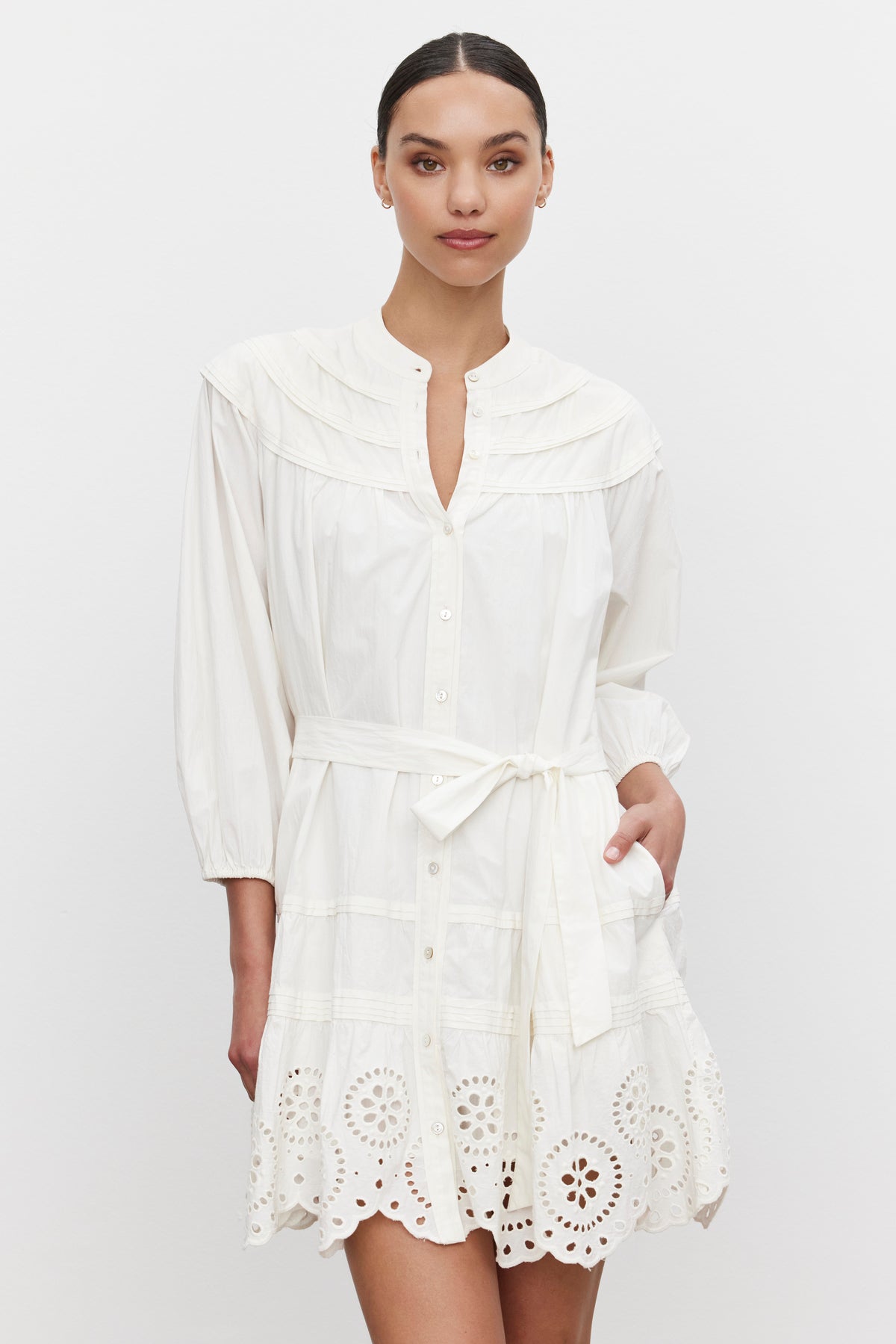   A person is wearing the BARB DRESS by Velvet by Graham & Spencer, a white, long-sleeved cotton dress with a belted waist and eyelet lace detailing at the hem. The dress also features a pleated skirt for a customizable fit. They are standing against a plain white background. 