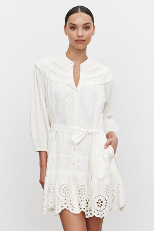 A person is wearing the BARB DRESS by Velvet by Graham & Spencer, a white, long-sleeved cotton dress with a belted waist and eyelet lace detailing at the hem. The dress also features a pleated skirt for a customizable fit. They are standing against a plain white background.