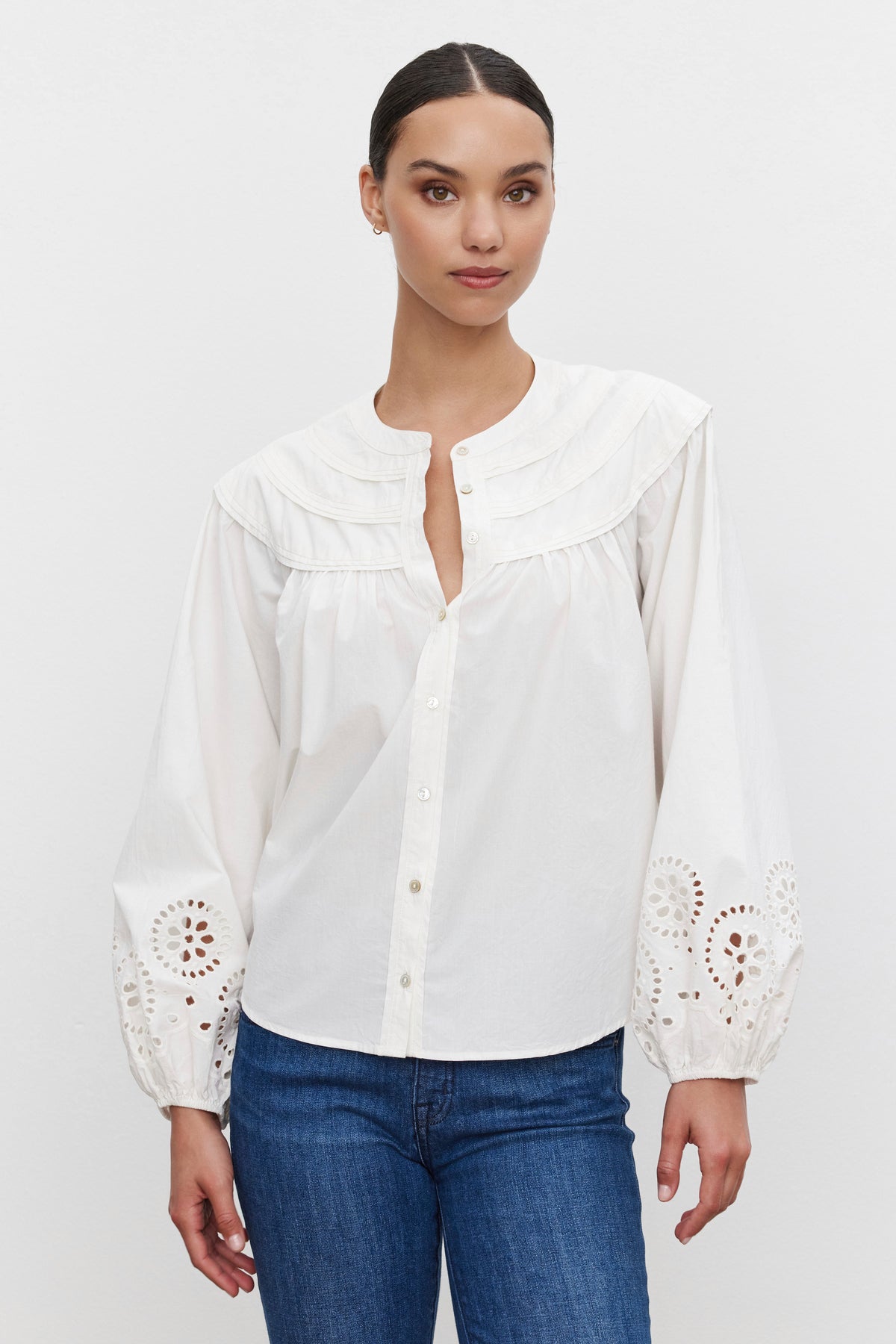   A person wearing the CARDEN TOP by Velvet by Graham & Spencer, a white cotton blouse featuring lace details on the sleeves, is paired with blue jeans and stands against a plain background, highlighting its timeless style with a banded collar and relaxed-fit design. 