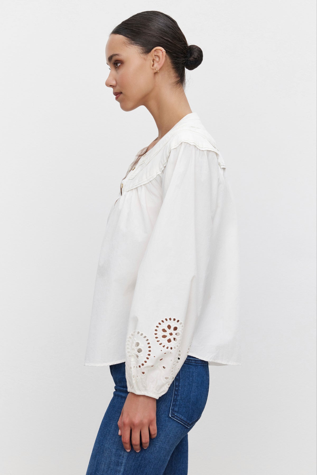   A person wearing the CARDEN TOP by Velvet by Graham & Spencer, featuring a relaxed fit and embroidered sleeves, along with blue jeans, stands in profile against a plain background. 