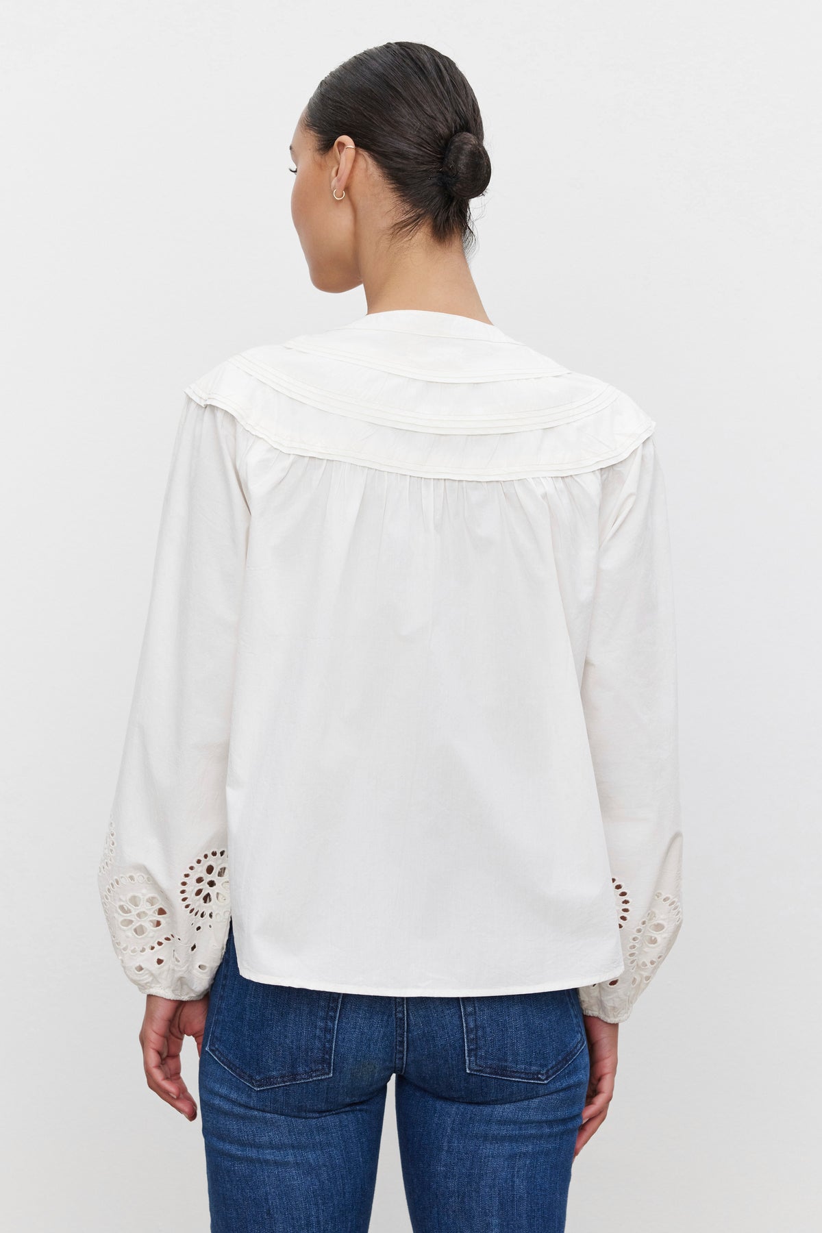   A person with dark hair in a bun is wearing the CARDEN TOP by Velvet by Graham & Spencer, a white cotton top featuring eyelet sleeves, along with blue jeans, and is facing away from the camera. 