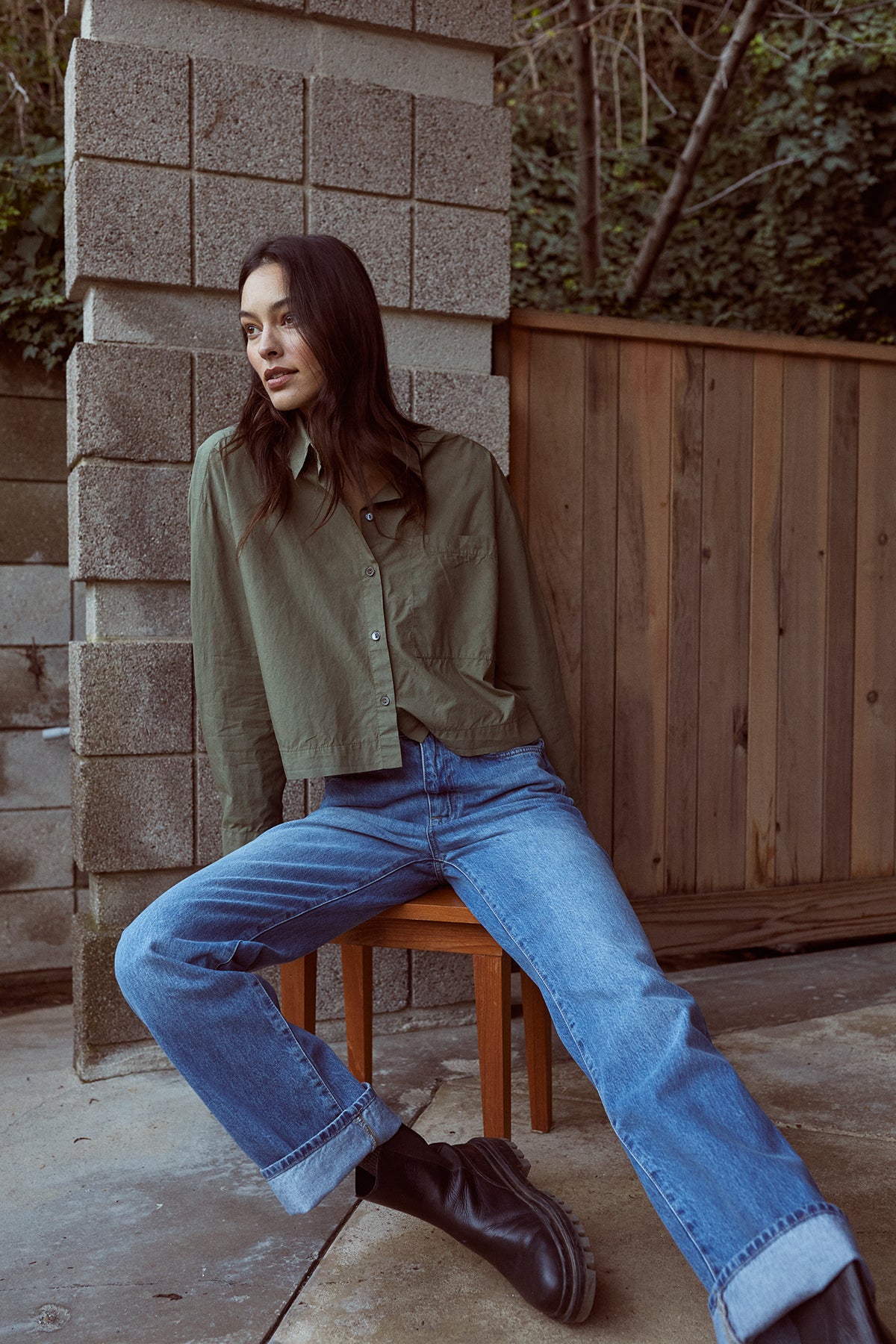   A woman is sitting on a chair wearing LUCILLE CROPPED BUTTON-UP SHIRT by Velvet by Graham & Spencer jeans and boots. 