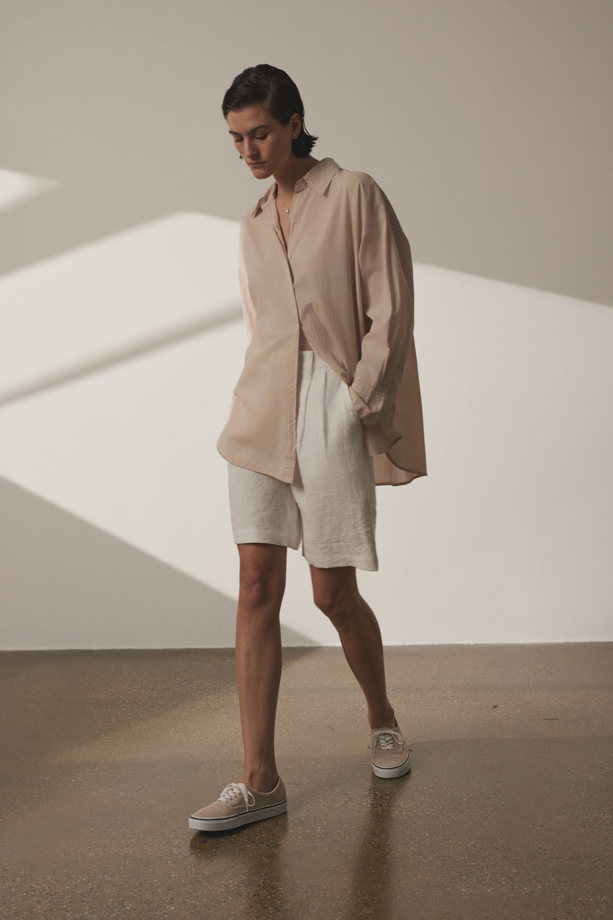 A person stands indoors, wearing an oversized Velvet by Jenny Graham REDONDO button-up shirt, white shorts, and tan sneakers, looking down thoughtfully in a sunlit room with shadows on the wall.-36863309185217