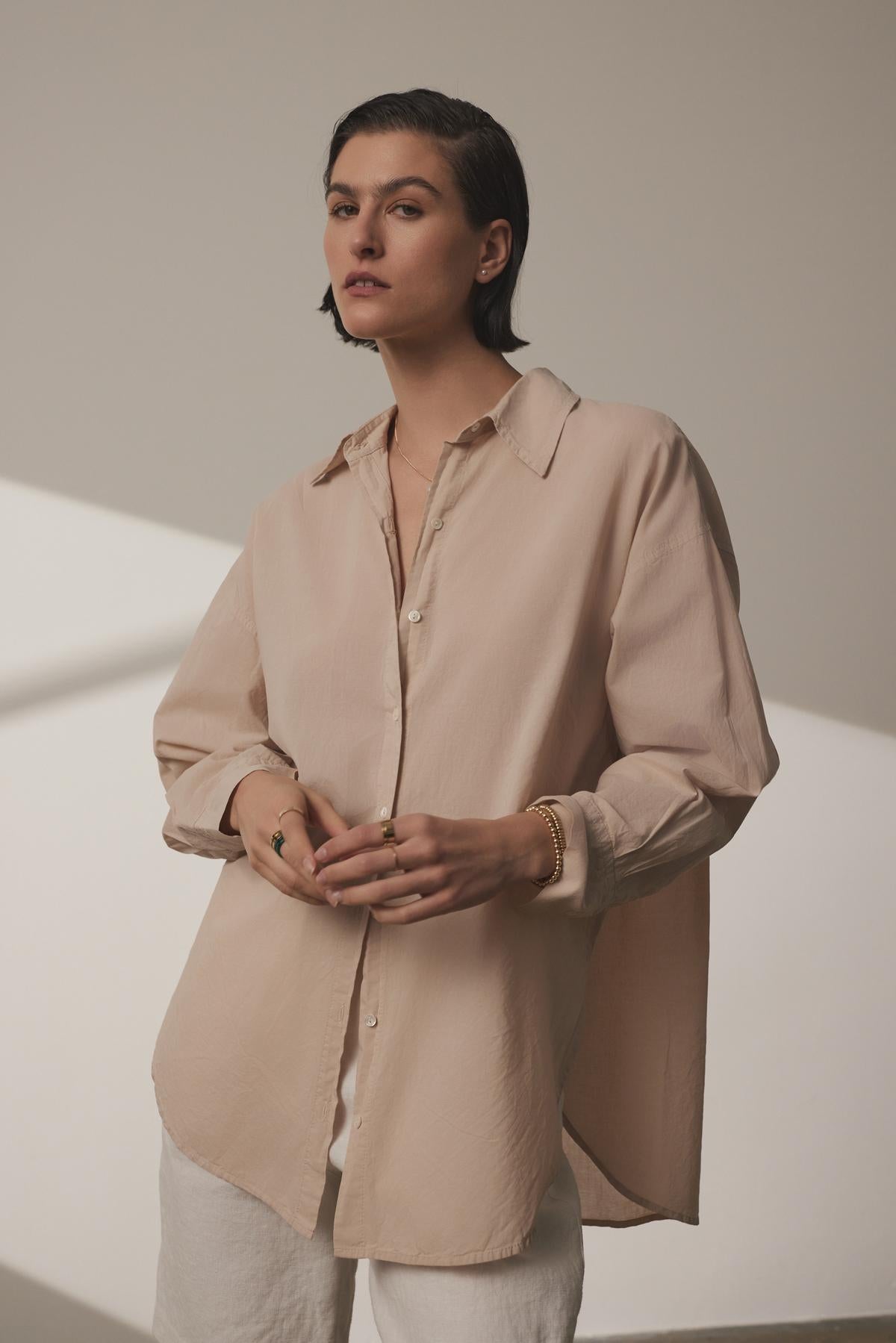 A woman in a Velvet by Jenny Graham REDONDO BUTTON-UP SHIRT and white relaxed trousers, standing with her hands clasped in front of her, against a softly lit beige background.-36863309152449