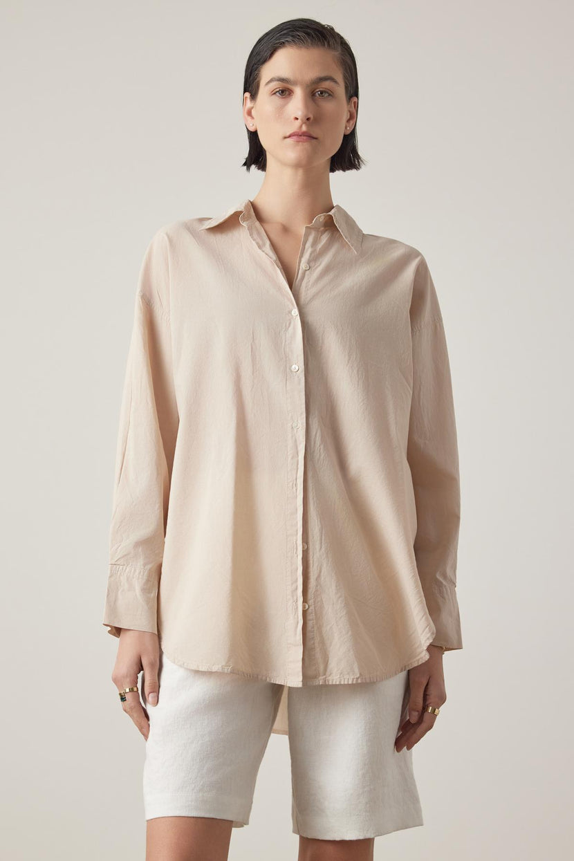 A person wearing the oversized REDONDO BUTTON-UP SHIRT in beige, made of soft cotton by Velvet by Jenny Graham, paired with white shorts, stands against a plain background, looking at the camera.