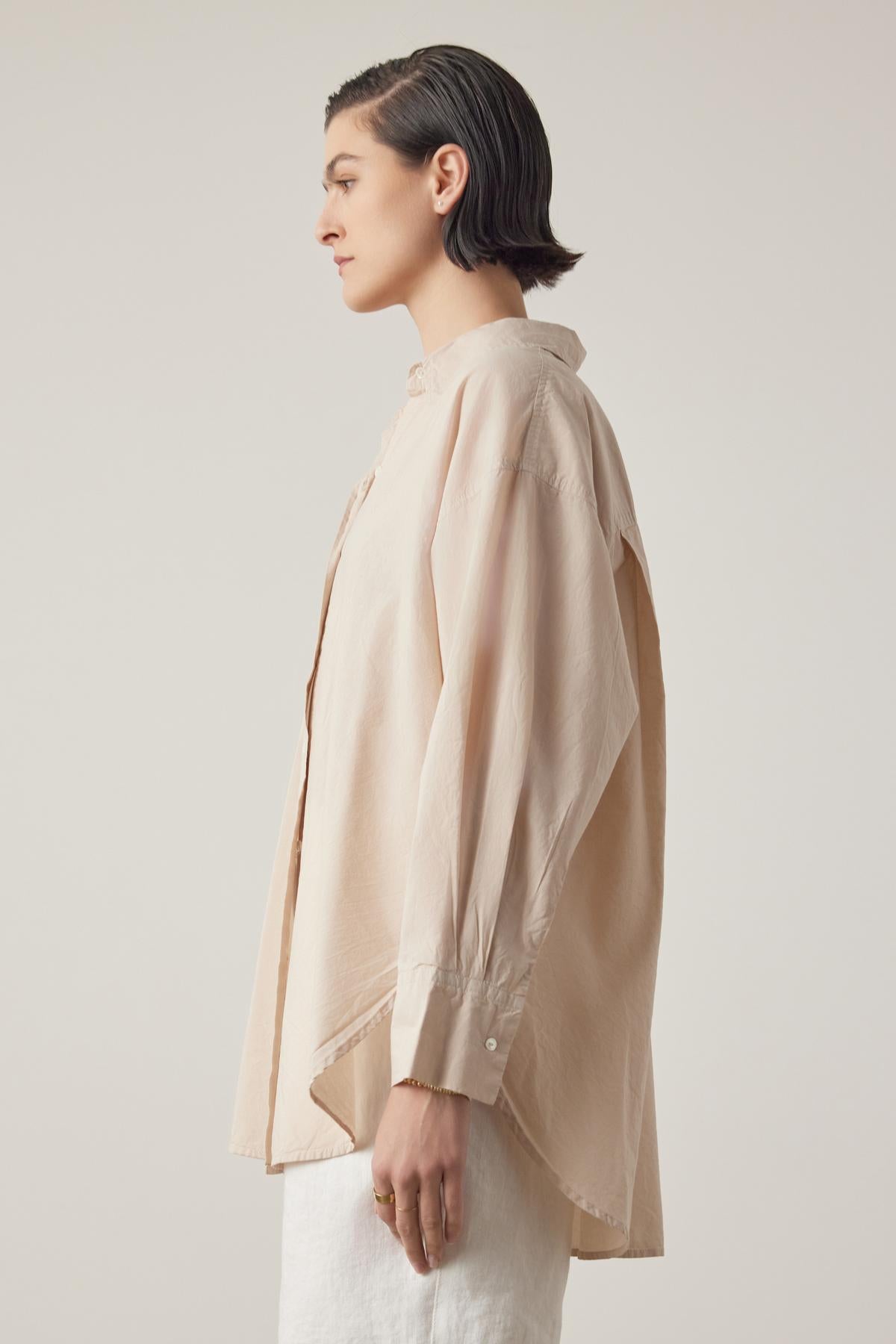   A person with short dark hair wears the oversized, light beige REDONDO BUTTON-UP SHIRT by Velvet by Jenny Graham and relaxed trousers, viewed from the side against a plain background. 